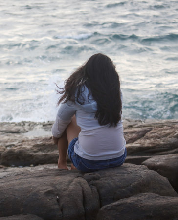 A woman sits on a rock by the ocean, looking out at the waves. The scene is peaceful and calming, with the sound of the waves providing a soothing background noise. The woman is lost in thoughtの写真素材