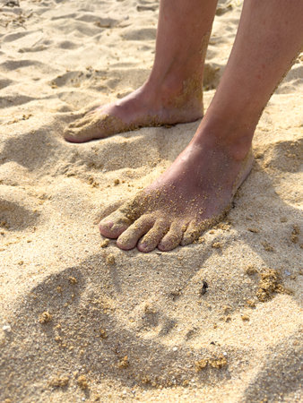 A person's feet are in the sand on a beach. The sand is light brown and the person's feet are...