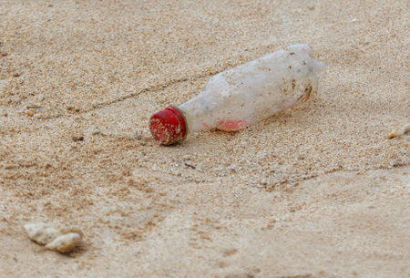 A bottle of water is laying on the sand. The bottle is empty and has a red cap. The bottle is on the beach and is surrounded by sandの写真素材