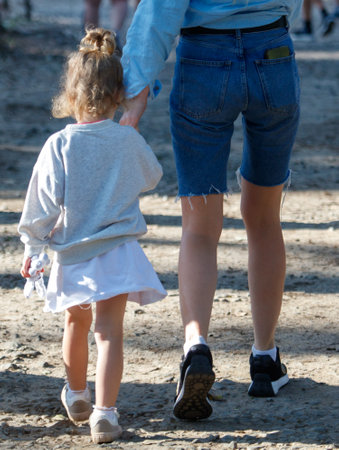 A woman is holding a child's hand while they walk on a dirt path. The child is wearing a white dress and the woman is wearing blue jeansの写真素材