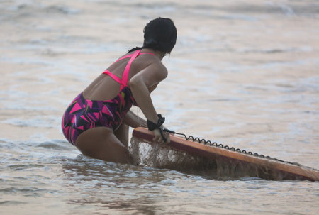 A woman in a pink and purple swimsuit is holding a surfboard in the water. She is bending over to pick it upの写真素材