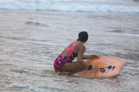 A woman in a pink and black swimsuit is holding an orange surfboard. She is kneeling in the water, preparing to go surfingの写真素材