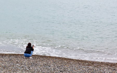 A woman is sitting on the beach, taking a picture of the ocean. Scene is peaceful and serene, as the woman is enjoying the beauty of the ocean and capturing it in a photographの写真素材