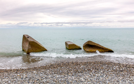 Three large rocks are sitting in the water near the shore. The water is calm and the sky is cloudyの写真素材