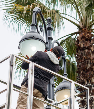 A man is fixing a street light on a pole. The light is on and the man is wearing a black jacketの写真素材