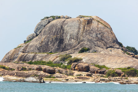 A rocky hill with a few trees on it. The hill is very large and the trees are sparse. The sky is clear and the water is calmの写真素材