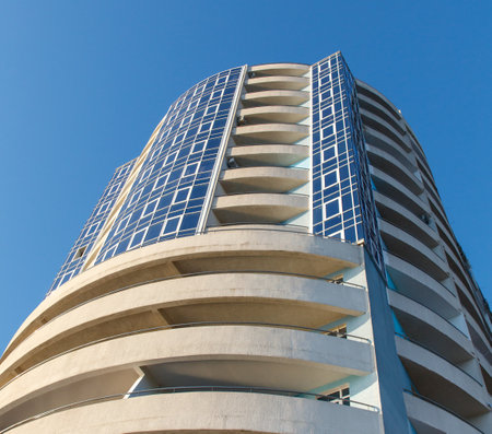 A tall building with a lot of windows and a balcony. The building is white and has a modern design. The sky is blue and clear, and the sun is shining brightlyの写真素材