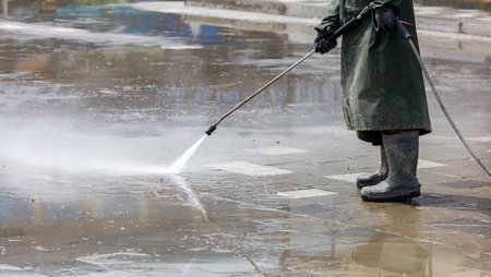 A man in a green coat is standing in the rain, holding a pressure washer. The scene is wet and gloomy, with the man's coat and boots getting soaked. The man is focused on his taskの写真素材