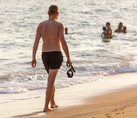 A man is walking on the beach with a snorkel in his hand. The beach is crowded with people, some of whom are swimming in the water. The scene is lively and full of energyの写真素材