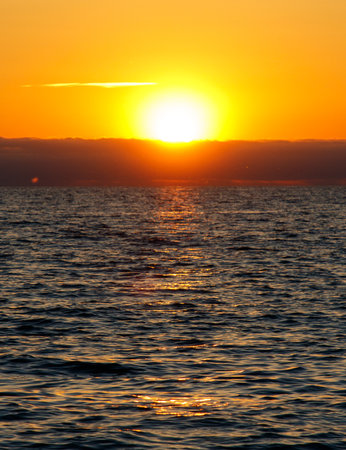 The sun is setting over the ocean, casting a warm glow on the water. The sky is a mix of orange and pink hues, creating a serene and peaceful atmosphere. The water is calm and stillの写真素材