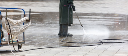 A man in a green coat is standing in the rain, holding a pressure washer. The scene is wet and gloomy, with the man's coat and boots getting soaked. The man is focused on his taskの写真素材
