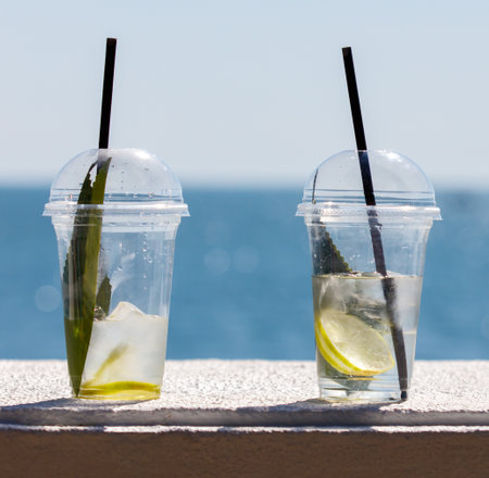 Two plastic cups with straws in them, one with a lime wedge in it. The cups are on a ledge near the oceanの写真素材