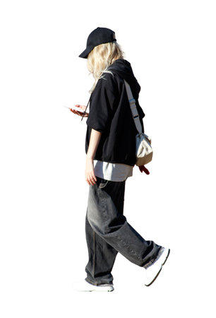 A woman in a black jacket and jeans is walking down the street while holding a cell phone. She is wearing a black hat and a white shirtの写真素材