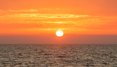 The sun is setting over the ocean, casting a warm glow on the water. The sky is filled with clouds, creating a serene and peaceful atmosphereの写真素材