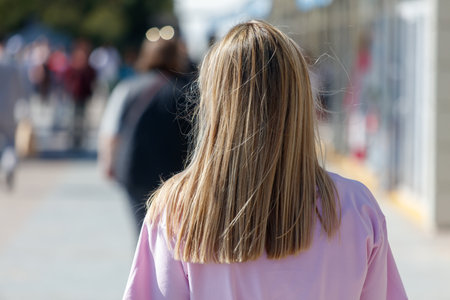 A woman with long blonde hair is walking down a sidewalk. She is wearing a pink shirt and has a backpack on her back. There are other people walking around herの写真素材