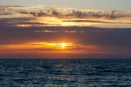 The sky is a beautiful orange color as the sun sets over the ocean. The water is calm and the horizon is visibleの写真素材