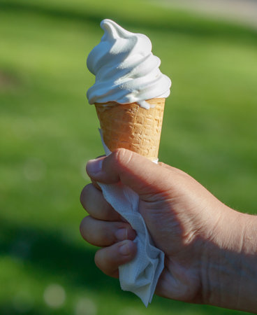 A person is holding a white ice cream cone with a napkin. The person is holding the cone with their hand, and it is a sunny day outsideの写真素材