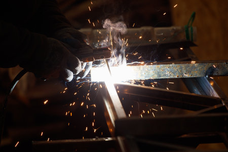 A man is welding a piece of metal. The sparks from the welding are flying everywhere, creating an intense atmosphere. The man is wearing gloves to protect his hands from the heatの写真素材