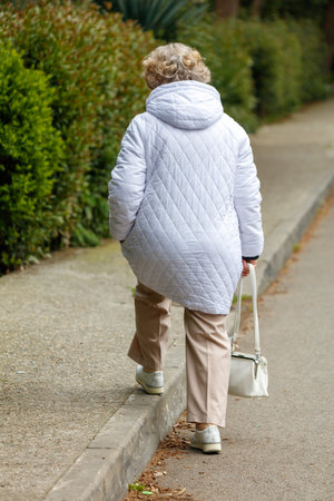 A woman in a white jacket and tan pants is walking down a sidewalk. She is carrying a white purse and a white jacketの写真素材