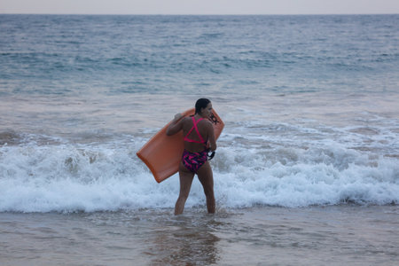 A woman is walking in the ocean with a surfboard. She is wearing a pink and black bikiniの写真素材