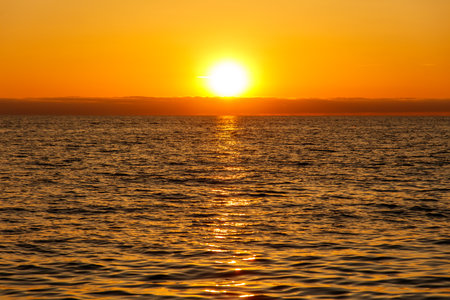 The sun is setting over the ocean, casting a warm glow on the water. The sky is a mix of orange and pink hues, creating a serene and peaceful atmosphere. The water is calm and stillの写真素材