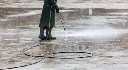 A man in a green coat is standing in the rain, holding a pressure washer. The scene is wet and gloomy, with the man's coat and boots getting soaked. The man is focused on his taskの写真素材