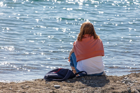 A woman sits on the beach with her back to the water. She is wearing a red scarf and a white shirt. The water is calm and the sky is clear. The scene is peaceful and relaxingの写真素材