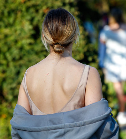 A woman with a ponytail and a backless dress is standing in a park. She is wearing a gray jacket and a white shirtの写真素材