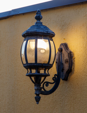 A lighted lamp post with a yellow wall behind it. The light is on and the lamp post is lit upの写真素材