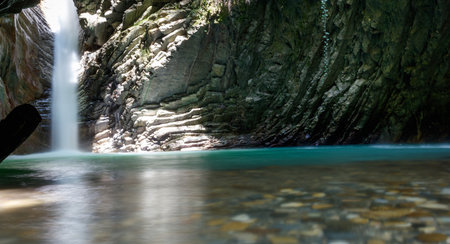 A waterfall is reflected in the water of a river. The water is clear and calm. The rocks surrounding the river are jagged and roughの写真素材
