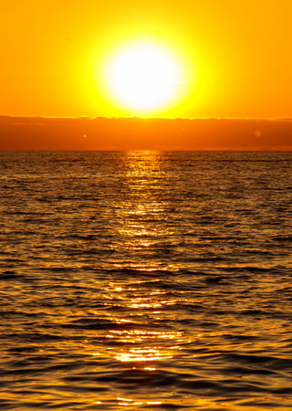 The sun is setting over the ocean, casting a warm glow on the water. The sky is a mix of orange and pink hues, creating a serene and peaceful atmosphere. The water is calm and stillの写真素材