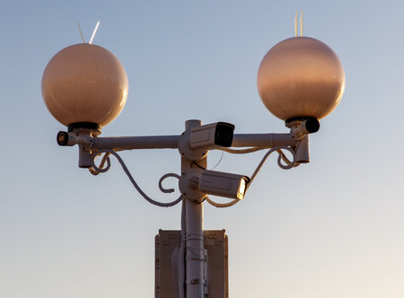 Two street lights with cameras on them. The street lights are white and the sky is blueの写真素材