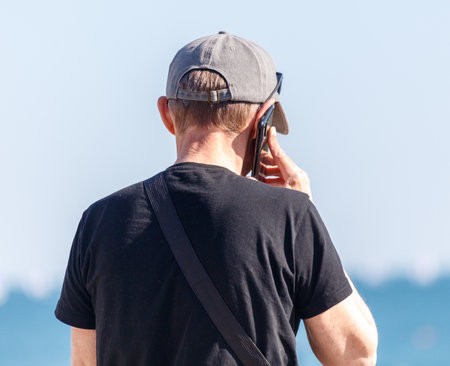 A man is talking on his cell phone while wearing a hat and a black shirt. He is walking on a beachの写真素材