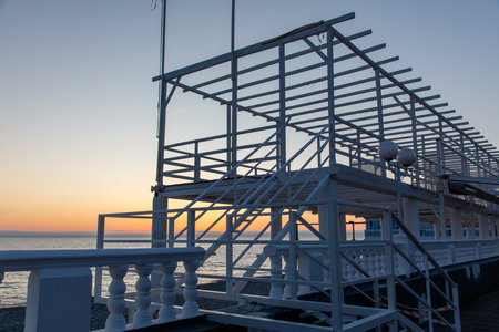 A pier with a white structure and a railing. The sky is orange and the water is blue