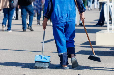 A man in a blue jumpsuit is walking down the street, holding a broom and a dustpan. He is likely a street cleaner or a janitor, as he is carrying the necessary tools to clean the areaの写真素材