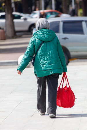 A woman wearing a green jacket and gray hat walks down a sidewalk with a red bag. The scene is set in a city with cars parked along the street. The woman is in a hurryの写真素材