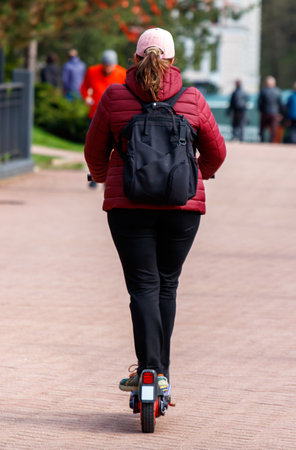A woman is riding a scooter down a sidewalk. She is wearing a red jacket and a pink hatの写真素材
