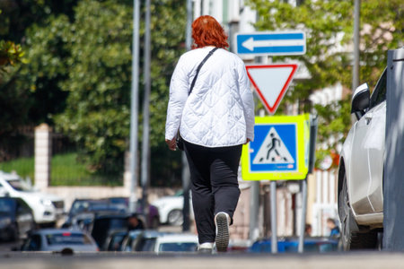 A woman walks down a street with a crosswalk sign in front of her. The street is busy with cars and other pedestriansの写真素材