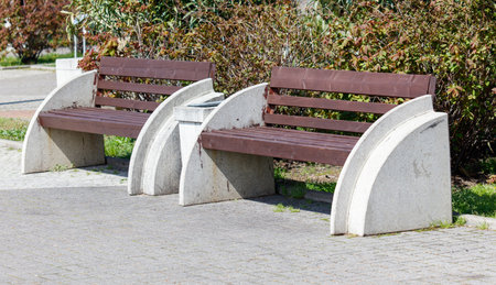 Two benches are sitting on a cement slab in front of a bush. The benches are brown and whiteの写真素材