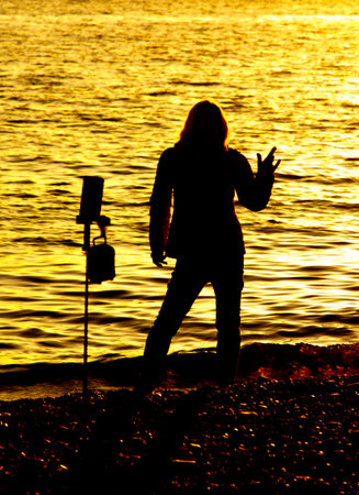 A woman stands on a beach at sunset, holding her hands up in the air. The scene is peaceful and serene, with the sun setting in the backgroundの写真素材