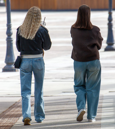 Two women walking down a sidewalk, one of them holding a cell phone. The other woman is wearing a black jacket and blue jeansの写真素材