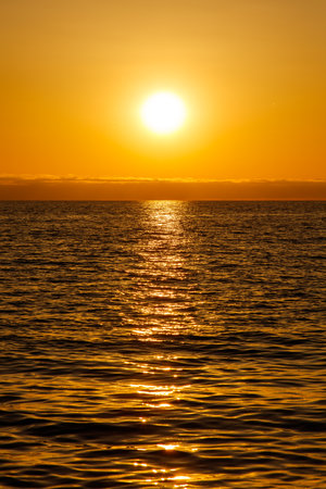 The sun is setting over the ocean, casting a warm glow on the water. The sky is a mix of orange and pink hues, creating a serene and peaceful atmosphere. The water is calm and stillの写真素材