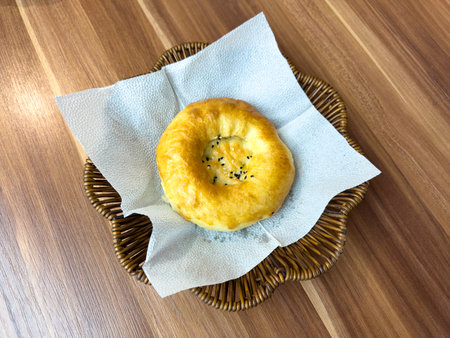 A small, round pastry sits on a napkin in a basket. The pastry is brown and has a sprinkle of sesame seeds on top. The basket is made of wicker and is placed on a wooden tableの写真素材