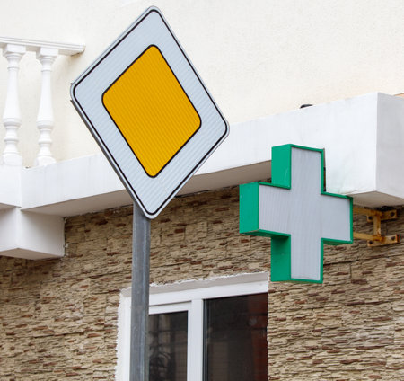 A street sign with a yellow square and a red arrow is leaning against a building. A green cross is hanging from the building next to the signの写真素材