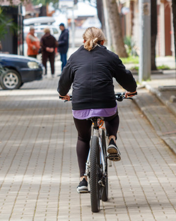 A woman is riding a bicycle down a street. The street is lined with cars and a few people are walking around. The woman is wearing a black jacket and a purple shirtの写真素材