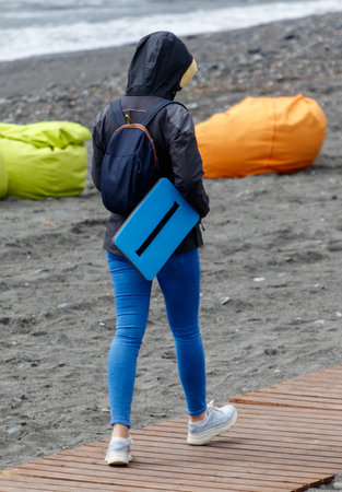 A woman is walking on a beach with a blue laptop bag. She is wearing a black jacket and blue jeansの写真素材
