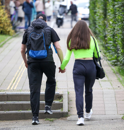 A man and a woman are walking down a sidewalk, holding hands. The man is wearing a backpack and the woman is wearing a green shirtの写真素材