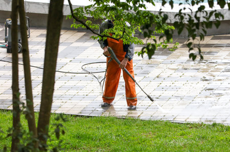 A man in orange jumpsuit is cleaning a sidewalk with a pressure washer. The scene is set in a park with trees in the backgroundの写真素材
