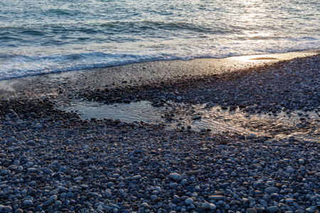 A rocky beach with a body of water in the background. The rocks are scattered all over the beach, and the water is calm. The scene has a peaceful and serene mood, with the sun shining on the waterの写真素材
