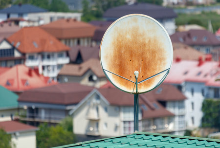 A rusty satellite dish is on top of a roof in front of a row of houses. The dish is old and worn, but it still serves its purpose. Concept of nostalgia and a connection to the pastの写真素材